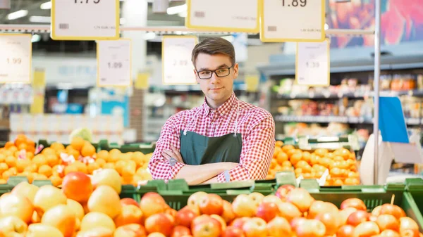 Au supermarché : Portrait du beau commis de stock portant un tablier, organisant des fruits et légumes biologiques, il sourit et croise les bras. Travailleur amical et efficace au magasin. — Photo