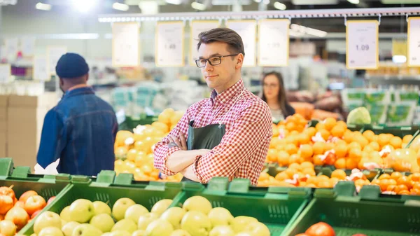 V supermarketu: Portrét pohledného skladového úředníka v zástěře, aranžmá organického ovoce a zeleniny, usmívá se a kříže zbraní. Friendly, Efficient Worker at the Store. — Stock fotografie