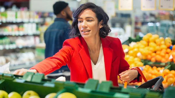 A szupermarketben: a gyönyörű mosolygó nő portréja organikus gyümölcsöket választ a Friss Termelés sorban, és kosárba teszi őket. — Stock Fotó