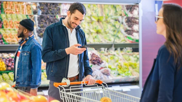 Στο σούπερ μάρκετ: Όμορφος άνθρωπος χρησιμοποιεί Smartphone, ενώ στέκεται στο Fresh Produce Τμήμα του καταστήματος. Άνθρωπος βυθισμένο στο Internet Surfing στο κινητό του τηλέφωνο στο παρασκήνιο πολύχρωμα φρούτα και — Φωτογραφία Αρχείου