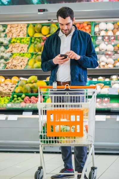 Au supermarché : bel homme avec smartphone, pousse le panier, marche dans la section des fruits et légumes frais du magasin. Homme plongé dans Internet Surfer sur son téléphone portable. — Photo