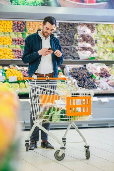 Im Supermarkt: Schöner Mann mit Smartphone, schiebt Einkaufswagen, geht an Frischeabteilung des Ladens vorbei. Mann mit Handy im Internet surft. — Stockfoto