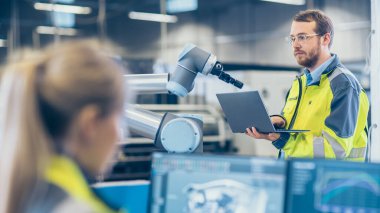 Fabrikada: Kadın Makine Mühendisi Kişisel Bilgisayarında 3D Motor Tasarımı Yaptı, Erkek Otomasyon Mühendisi Robotik Kolu Programlamak için Dizüstü Bilgisayarı Kullandı.
