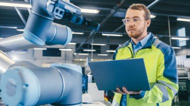 Fabrikada Düşük Açı Atışı: Otomasyon Mühendisi Robot Kolu Denemek ve Programlamak için Dizüstü Bilgisayar Kullanıyor. Otomatik Üretim Endüstrisinde Yeni Dönem.