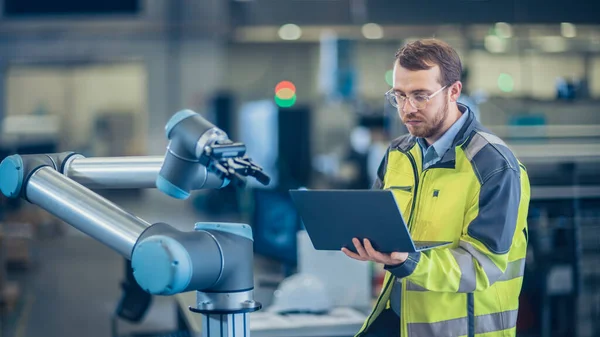 En la fábrica: El ingeniero de automatización utiliza el ordenador portátil para programar el brazo robótico. Nueva era en la industria manufacturera automática. —  Fotos de Stock