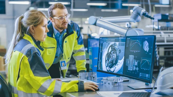 En la fábrica: Ingeniero mecánico masculino e ingeniero jefe femenino trabajan juntos en la computadora personal, discuten detalles del modelo de motor 3D para brazo robótico. — Foto de Stock