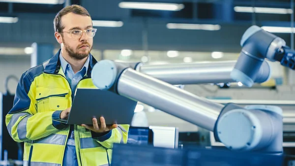 À l'usine : l'ingénieur en automatisation utilise un ordinateur portable pour programmer un bras robotique. Nouvelle ère dans l'industrie manufacturière automatique. — Photo