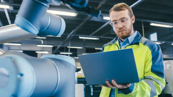 En la fábrica: El ingeniero de automatización utiliza el ordenador portátil para programar el brazo robótico. Nueva era en la industria manufacturera automática. — Foto de Stock