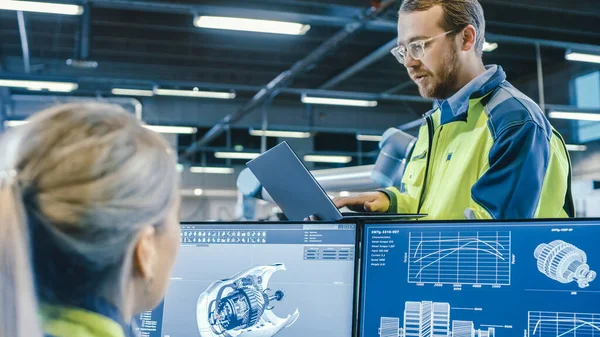 På fabriken: Man och kvinna mekaniska ingenjörer använder laptop och persondator när du diskuterar Robotic Arm Manufacturing Project. Skärmen visar 3D Engine Concept tillverkad med CAD-programvara. — Stockfoto