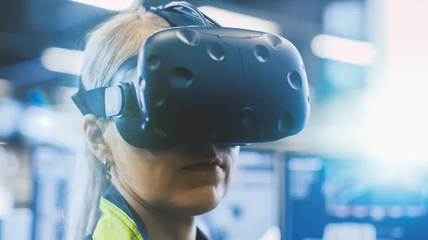 En la fábrica: La ingeniera femenina que usa auriculares de realidad virtual trabaja en un proyecto, moviendo su cabeza. En las pantallas de fondo y manufactura industrial. —  Fotos de Stock