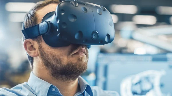 Porträtaufnahme des Wirtschaftsingenieurs, der Virtual-Reality-Headset trägt und mit Controllern gestikuliert. Im Hintergrund Displays und Produktionsanlagen aus dem Fokus. — Stockfoto