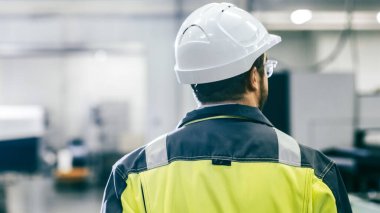 Arka planda Otomatik Makineler ile Modern Üretim Tesisinde Yürüyen Koruyucu Giysi Giyen Endüstriyel Mühendisin Arka Görünümü.