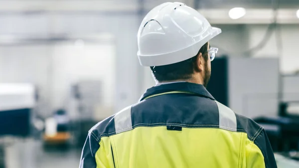 Vista posterior La toma del ingeniero industrial que usa ropa protectora camina a través de una instalación de fabricación moderna con maquinaria automática trabajando en segundo plano. — Foto de Stock