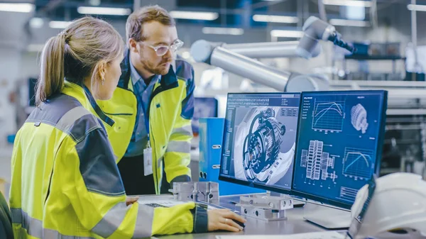 À l'usine : Un ingénieur mécanique masculin tient un composant tandis qu'une ingénieure en chef travaille sur un ordinateur personnel, ils discutent des détails de la conception du modèle de moteur 3D. — Photo