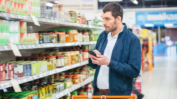 I snabbköpet: Stilig man använder smartphone och ser på näringsvärde av konserver varor. Hes Stående med kundvagn i Konserverade varor sektionen. — Stockfoto