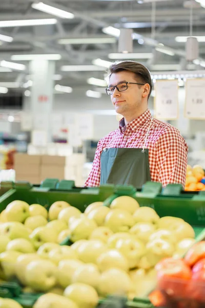 V supermarketu: Portrét Usmívajícího se skladového úředníka v zástěře, aranžmá organického ovoce a zeleniny. Friendly, Efficient Worker at the Farmers Market Section of the Store — Stock fotografie