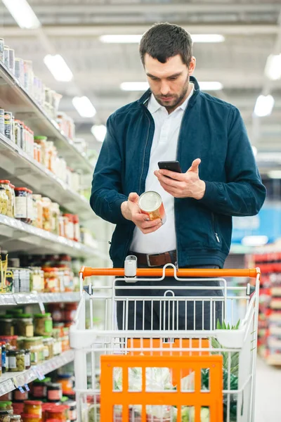 At the Supermarket: Handsome Man Uses Smartphone to Check Nutritional Value of the Canned Goods and Buy it. Hes Standing with Shopping Cart in Canned Goods Section. — Stock Photo, Image