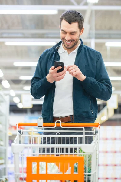No supermercado: Homem bonito usa smartphone e caminha através da prateleira de produtos enlatados. Hes Empurrando o carrinho de compras através da seção de produtos enlatados. — Fotografia de Stock