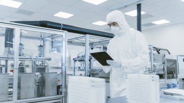 Kalite Kontrol: Dijital Tablet Bilgisayar kullanan ve koruyucu giyimli bilim adamı imalat laboratuarından geçiyor. Ürün Üretimi: İlaç, Yarı iletkenler, Biyoteknoloji.