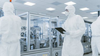 Kalite Kontrol: Dijital Tablet Bilgisayar kullanan ve koruyucu giyimli bilim adamı imalat laboratuarından geçiyor. Ürün Üretimi: İlaç, Yarı iletkenler, Biyoteknoloji.