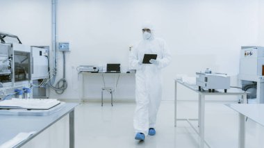 Kalite Kontrol: Dijital Tablet Bilgisayar kullanan ve koruyucu giyimli bilim adamı imalat laboratuarından geçiyor. Ürün Üretimi: İlaç, Yarı iletkenler, Biyoteknoloji.