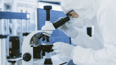 Koruyucu Giysiler Laboratuvarında Araştırma Yapan Bilimadamı Mikroskop ve Kişisel Bilgisayar kullanır. Modern Manufactory Yarı iletkenler ve İlaç Ürünleri Üretimi.