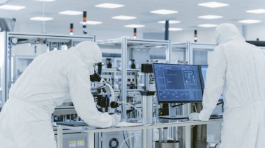 Koruyucu Giysiler Laboratuvarında Araştırma Yapan, Mikroskop kullanan ve Kişisel Bilgisayara Veri Giren Bilimadamları. Modern Manufactory Yarı iletkenler ve İlaç Ürünleri Üretimi.