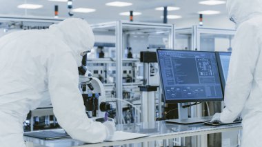 Koruyucu Giysiler Laboratuvarında Araştırma Yapan, Mikroskop kullanan ve Kişisel Bilgisayara Veri Giren Bilimadamları. Modern Manufactory Yarı iletkenler ve İlaç Ürünleri Üretimi.