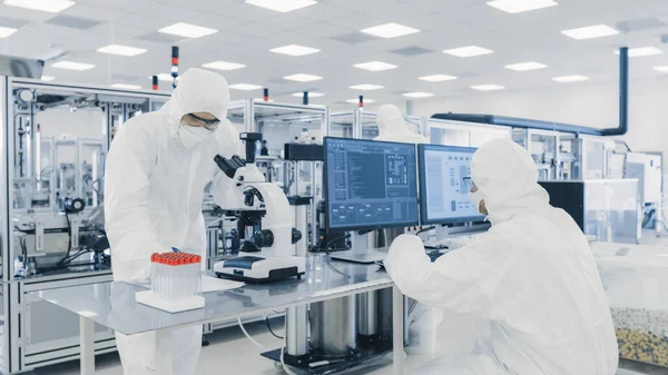 Kutatók csoportja steril öltönyben Számítógépekkel dolgozik, mikroszkóp alatt néz és modern ipari gépekkel dolgozik a laboratóriumban. Termékgyártási folyamat: Gyógyszeripar — Stock Fotó