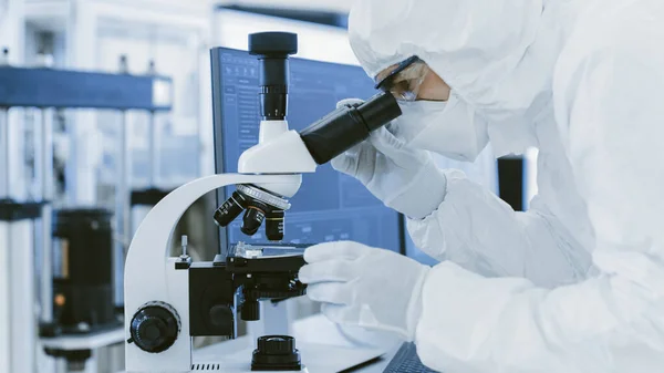 У лабораторії в захисному одязі використовує мікроскоп і персональний комп'ютер. Сучасне виробництво напівпровідників та фармацевтичних продуктів . — стокове фото