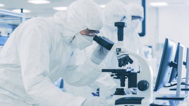 Koruyucu Giysiler Laboratuvarında Araştırma Yapan, Mikroskop kullanan ve Kişisel Bilgisayara Veri Giren Bilimadamları. Modern Manufactory Yarı iletkenler ve İlaç Ürünleri Üretimi.