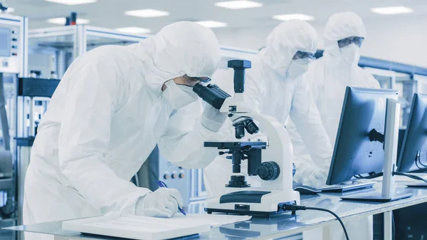 在《穿着防护服做研究的实验室科学家》中，使用显微镜并将数据写入笔记本。现代手工生产半导体和药品. — 图库照片