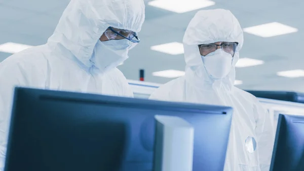 Dos científicos técnicos en trajes estériles comprueban los datos en el ordenador personal, la tienen discusión. Trabajan en el Laboratorio de Fabricación de Pharmateutics. —  Fotos de Stock