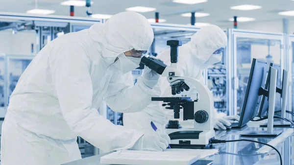 Em Laboratório Cientista em Roupas de Proteção Fazendo Pesquisa, Usando Microscópio e Escrevendo Dados. Trabalhadores que trabalham em uma manufatura moderna produzindo semicondutores e itens farmacêuticos. — Fotografia de Stock