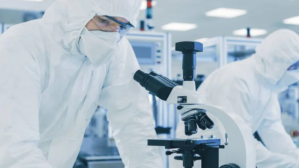 In laboratorio Scienziato in indumenti protettivi facendo ricerca, utilizzando microscopio e annotare i dati. Lavoratori che lavorano su una manifattura moderna che produce semiconduttori e articoli farmaceutici. — Foto Stock