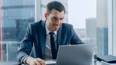 Başarılı bir iş adamı masasında oturuyor, bilgisayarını kullanıyor. Pencere Büyük Şehir İş Bölgesi Manzarasında.