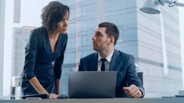 Ofisin kadın CEO 'su masasında oturan önde gelen iş adamıyla konuşuyor, dizüstü bilgisayar kullanıyor. Devam eden iş sözleşmelerini ve anlaşmaları tartışıyorlar. Arkaplanda Büyük Şehir Görünümü.