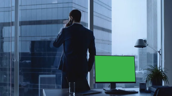 Empresário confiante em seu escritório fazendo chamada telefônica, computador pessoal em sua mesa mostra tela chave Chroma verde. Na janela Panoramic Big City View. — Fotografia de Stock