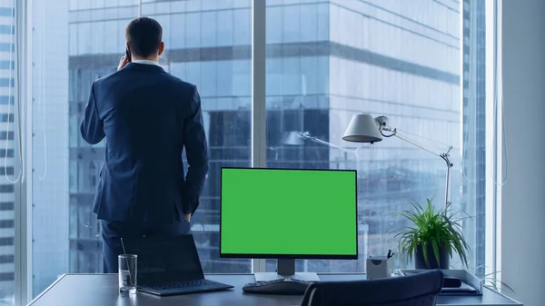 Selbstbewusster Geschäftsmann in seinem Büro telefoniert, Personal Computer auf seinem Schreibtisch zeigt grünen Chroma-Schlüsselbildschirm. Im Fenster Großstadtblick. — Stockfoto