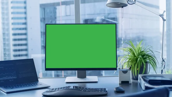 在写字楼的写字台上放了绿色模拟屏幕个人电脑。具有大城市商务区视野的现代风格房间. — 图库照片