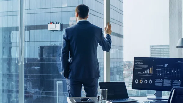 Skjuten av den självsäkra affärsmannen i en kostym stående i Office och tittar ut genom fönstret tankeväckande. Snygg modern Business Office med persondator och storstadsutsikt. — Stockfoto