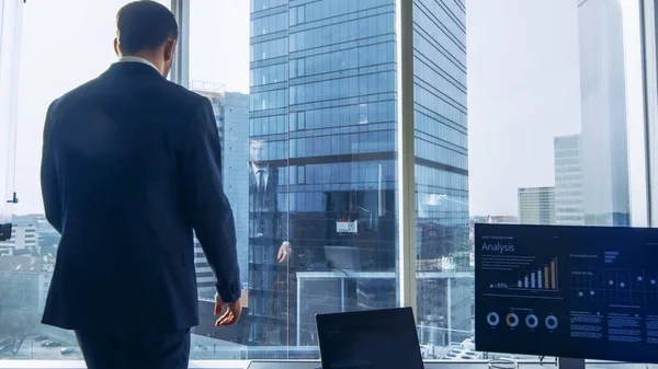Egy öltönyös üzletemberről, aki átsétál az irodáján, és figyelmesen néz ki az ablakon. Stílusos Modern Üzleti Iroda személyi számítógép és a Big City View. — Stock Fotó