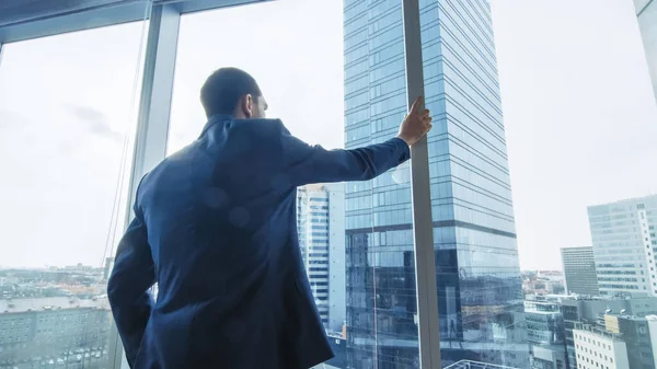 Thoughtful Businessman Wearing Suit Standing in His Office, Looking out of the Window and Contemplating Next Big Business Contract. Major City Business District with Panoramic Window View. Blue Colors — Stock Photo, Image