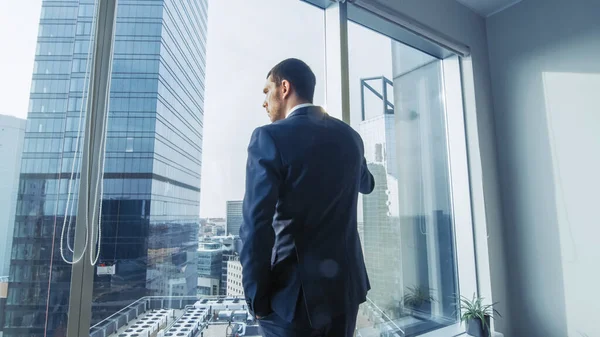 スーツを身に着けている思考のビジネスマンは彼のオフィスに立ち、窓の外を見、次の大きいビジネス契約を熟考する。パノラマウィンドウビューを備えた主要都市ビジネス地区。青い色 — ストック写真