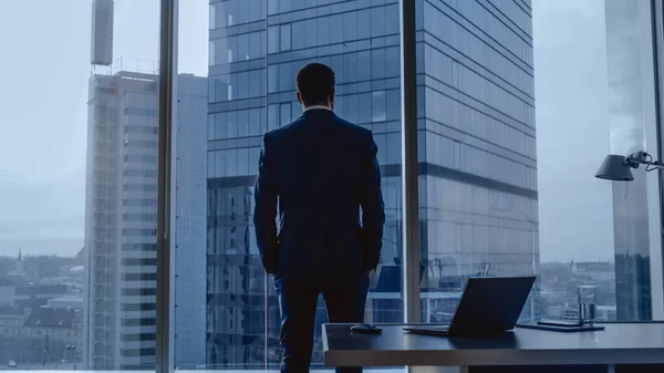 Achteraanzicht van de bedachtzame zakenman met een pak in zijn kantoor, handen in de zakken kijkend uit het raam. Big City Business District Panoramisch uitzicht op het venster. Donker regenweer. — Stockfoto