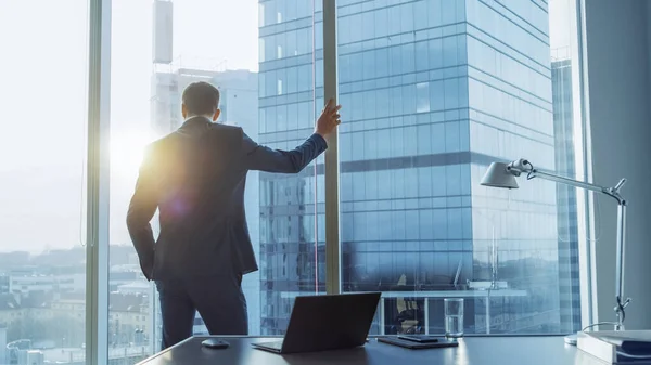 Skjuten av den självsäkra affärsmannen i kostym i sitt kontor lutande mot fönsterramen, tittar ut genom fönstret tankeväckande. Storstadens affärsdistrikt panoramafönster Visa. — Stockfoto