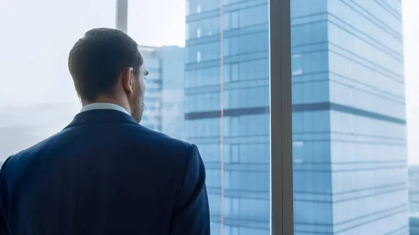 Medium Shot of Successful Businessman iført en dress stående på kontoret sitt, Contemplating Next Big Business Deal, ser ut av vinduet. Vinduet i storbyområdet Panoramautsikt. – stockfoto