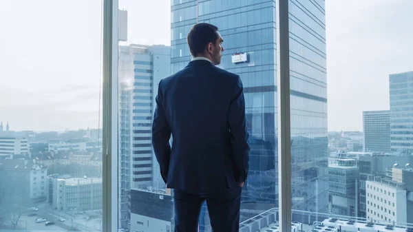 Skott av självsäker affärsman i kostym i sitt kontor, sätta händerna i fickor och titta ut genom fönstret tankeväckande, Fundera nya affärer och framtida kontrakt. — Stockfoto