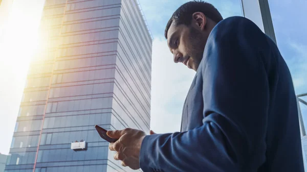 Χαμηλή γωνία Shot του επιτυχημένου επιχειρηματία σε ένα κοστούμι στέκεται στο γραφείο του, χρησιμοποιώντας το κινητό τηλέφωνο, ενώ στέκεται δίπλα στο παράθυρο με θέα στη μεγάλη πόλη. — Φωτογραφία Αρχείου