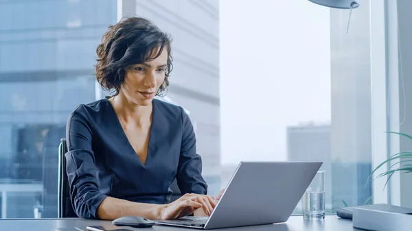 Empresaria segura sentada en su escritorio y trabajando en una computadora portátil en su oficina moderna. Elegante hermosa mujer haciendo trabajo importante. En la ventana Big City Business District View. —  Fotos de Stock